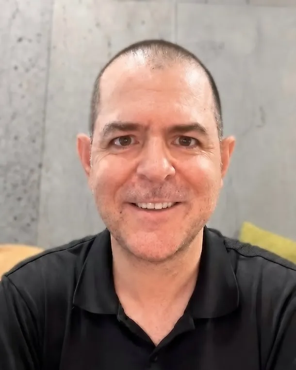 Jacob Hite, smiling at the camera in a black polo shirt, seated against a concrete wall.