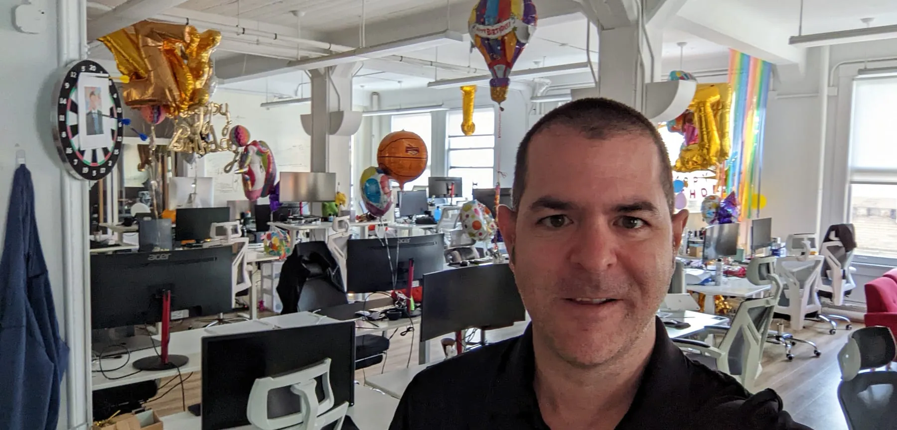 Jacob Hite in an open-plan engineering office, rows of desks and monitors behind him and birthday balloons hanging from the ceiling.