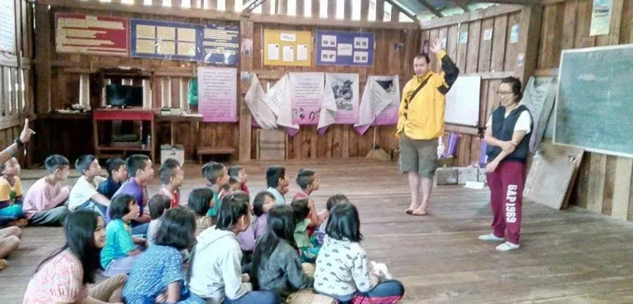 A room of children seated on the floor of a wooden schoolhouse in South East Asia, with two adults teaching at the front.
